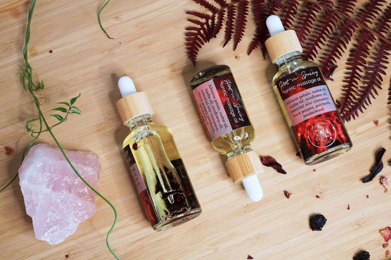 Drei Glasflaschen mit Pipette für "Hot n Spicy Tantric Massage Oil" sind diagonal auf einer hellen Holzoberfläche angeordnet. Eine Flasche liegt auf der Seite und zeigt botanische Zutaten, die im Öl enthalten sind. Verstreute getrocknete Blütenblätter, eine grüne Ranke, ein rosa Rosenquarzkristall und ein roter Farn schmücken die Szene.