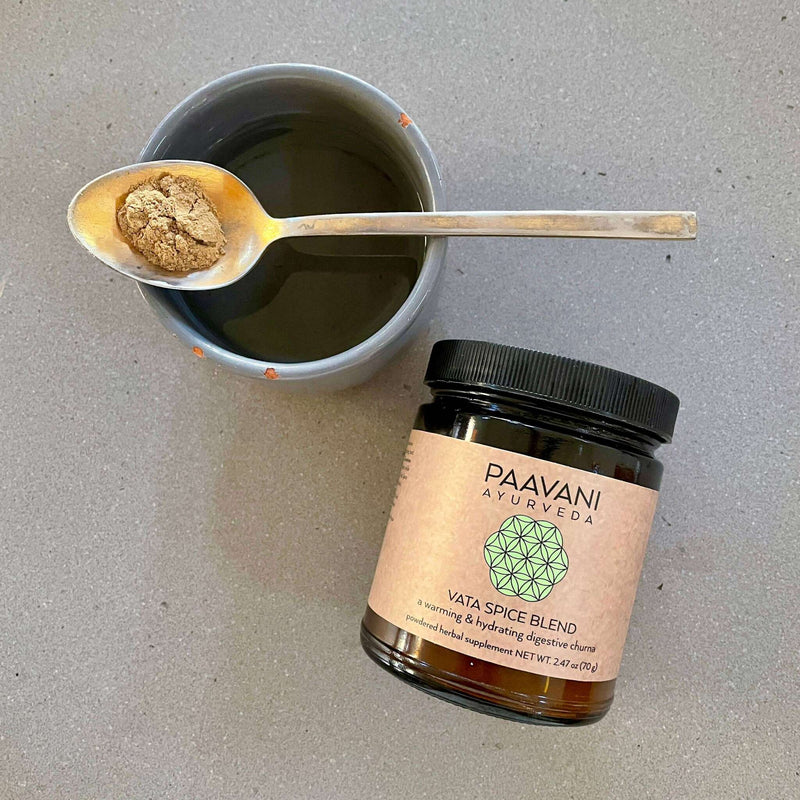  Eine Tasse mit dunkler Flüssigkeit und einem Löffel, der hellbraunes Pulver enthält, steht neben einem Glas "PAAVANI AYURVEDA VATA SPICE BLEND" auf einer grauen Oberfläche, was auf die Zubereitung eines Getränks hindeutet.