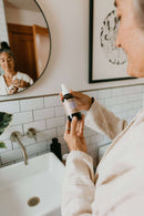 Eine Frau im Bademantel hält eine Flasche Paavani tridoshic Gesichtstoner über einem Waschbecken, während ihr Spiegelbild im runden Spiegel im Hintergrund zu sehen ist.