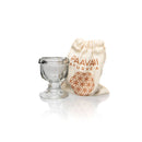  Ein transparentes Glas-Augenbad mit einem Paavani-gebrandeten Beutel aus naturfarbenem Stoff, der das gleiche geometrische Muster wie die Marke aufweist, auf einem weißen, reflektierenden Hintergrund.