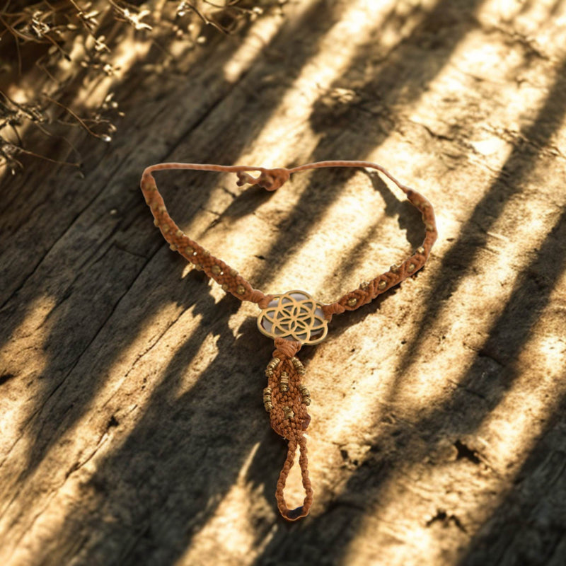 Ein handgefertigtes, braunes Makramee-Armband mit einem goldenen, filigranen Blumen des Lebens-Anhänger liegt auf einer Holzoberfläche mit Sonnenlicht und Schatten.