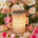 Eine beleuchtete, keramische Duftlampe/Wärmediffusor mit einem ausgeschnittenen Equalizer-Muster steht auf einer Oberfläche, umgeben von unscharfen rosa und weißen Blüten.