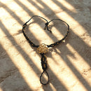  Ein schwarzes Makramee-Armband mit einem goldenen "Blume des Lebens"-Anhänger und kleinen goldenen Perlen liegt auf einem hellbraunen, steinernen Untergrund mit starken Schattenstreifen.