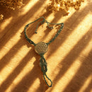 Ein grünes Makramee-Armband mit einem runden, goldenen Anhänger, der ein geometrisches Muster zeigt, liegt auf einem holzähnlichen Untergrund mit verschwommenen goldenen Elementen im Hintergrund.