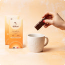 Eine Hand mit rotem Nagellack hält ein Stück Delikao "Get up & Go" Schokolade über einer weißen Tasse mit geprägtem Blumenmuster, auf einem beigefarbenen Hintergrund.