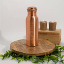 Eine Kupfer-Wasserflasche mit einer eingravierten Säule von Chakra-Symbolen, stehend auf einem runden Holzbrett mit zwei brennenden Kerzen.