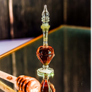 Eine kleine, verzierte Glasflasche mit klarem Stopfen, die ein dunkelbraunes Duftöl (Attar) enthält, reflektiert auf einer Spiegelfläche.