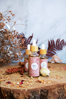 Drei Flaschen "Friya Skincare Oil" stehen auf einem Holzscheit, umgeben von getrockneten Blumen und Blättern. Der Hintergrund ist eine blaue Marmoroberfläche.