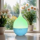 Ein elektrischer, tropfenförmiger Aroma-Diffusor mit blauem Boden und leuchtend grünem Oberteil, der Nebel ausstößt und auf einem Fensterbrett zwischen zwei Töpfen mit weißen Gänseblümchen steht.