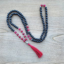 Eine lange Mala-Gebetskette aus polierten schwarzen und rosa Perlen, mit einem silbernen Buddha-Kopf und einer leuchtend roten Quaste, ausgelegt auf einer hellen Holzoberfläche.