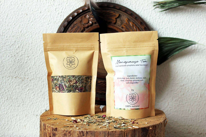 Zwei braune Standbodenbeutel "Menopause Tee" sind auf einem Baumstumpf aus Holz ausgestellt. Der vordere Beutel hat ein Sichtfenster, das die getrocknete Kräutermischung zeigt, und das Etikett des anderen Beutels enthält die Zutaten. Verstreute getrocknete Kräuter sind um die Basis der Beutel herum sichtbar, vor einer strukturierten weißen Wand mit einem geschnitzten Holzelement und grünen Palmblättern.