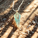 Eine Nahaufnahme eines goldenen Anhängers mit einem dreieckigen Opalstein, der auf einer strukturierten Oberfläche mit verstreuten Kristallen liegt, während sanftes Sonnenlicht Schatten wirft.