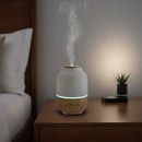Aroma-Diffusor aus Holz und weißer Keramik mit blauem Licht und aufsteigendem Dampf auf einem Nachttisch.
