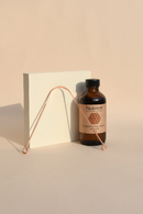 Eine braune PAAVANI Zimt-Ölziehe-Flasche steht neben einem weißen rechteckigen Block und einem kupferfarbenen Zungenreiniger, die lange Schatten auf einem hellen Pfirsich-Hintergrund werfen.