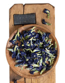 Eine Aufnahme von oben einer Holzschale, gefüllt mit getrockneten, blau-grünen Schmetterlingserbsenblüten (Clitoria ternatea), die auf einem rustikalen Holzbrett mit einem kleinen, schwarzen Metallgewicht liegt.