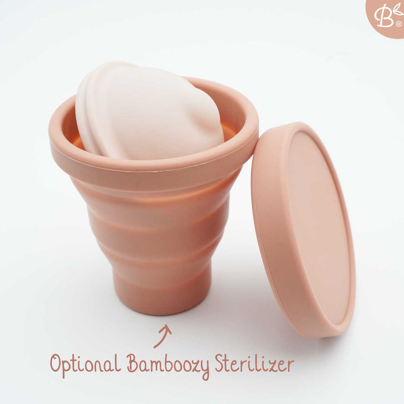 Eine gefaltete rosa Menstruationsscheibe in einem geöffneten, zusammenklappbaren Bamboozy Sterilisationsbecher, hebt das optionale Zubehör hervor.