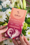 Ein Paar Hände hält eine Tafel delikao „Love Tonic Passion“ 71% Zartbitterschokolade mit Karamell und Passionsfrucht, angereichert mit Damiana, Maca und Zink. Die Verpackung mit pink-orangenem Farbverlauf wird vor einem weichen Hintergrund aus weißen und grünen Blumen präsentiert.