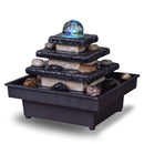 Ein mehrstufiger Tischbrunnen aus Stein mit einer leuchtenden Glaskugel und glatten Flusssteinen vor einem schlichten weißen Hintergrund.