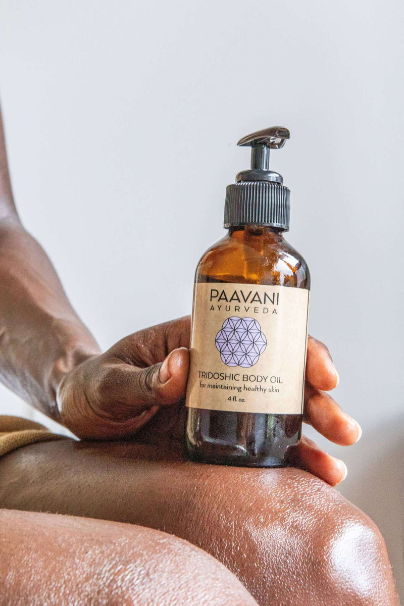 Eine glänzende, dunkle Haut, auf der ein Paavani Tridoshic Balancing Body Oil steht, das mit einer Hand gehalten wird.
