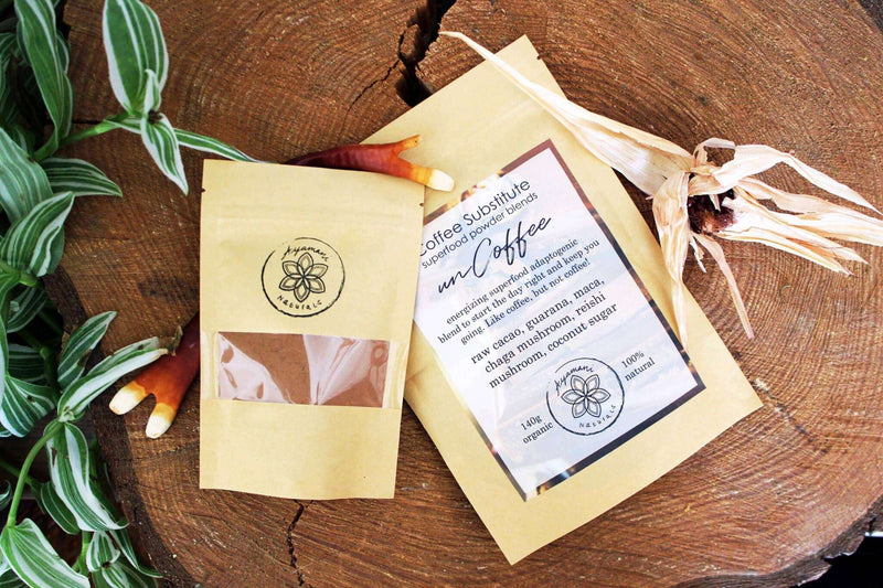 Zwei „Ayamanu Naturals Kaffee-Ersatz“-Superfood-Pulvermischungsbeutel werden auf einem großen, strukturierten Baumstamm präsentiert. Im Vordergrund befindet sich eine lebhafte grün-weiß gesprenkelte Pflanze, während getrocknete Maisblätter und getrocknete Reishi-Pilze (braun und orange) verstreut sind, was das natürliche und organische Thema unterstreicht.Zwei „Ayamanu Naturals Kaffee-Ersatz“-Superfood-Pulvermischungsbeutel werden auf einem großen, strukturierten Baumstamm präsentiert. Im Vordergrund befind