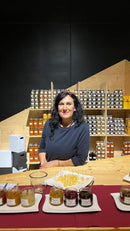 Eine Frau mit dunklem Haar steht hinter einer Holztheke und präsentiert verschiedene Honiggläser mit Proben auf Tellern. Die Regale hinter ihr sind mit ordentlich angeordneten Gläsern gefüllt, was eine einladende und handwerkliche Atmosphäre schafft.