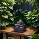 Ein mehrstufiger Tischbrunnen aus Stein mit einer leuchtenden Kristallkugel auf einem Holztisch, umgeben von üppigen grünen tropischen Pflanzen.