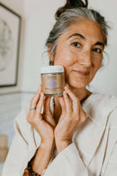 Eine lächelnde Frau mit salz- und pfefferfarbenem Haar in einem weiß gestreiften Oberteil hält einen durchsichtigen Tiegel mit PAAVANI "tridoshic" Reiniger und Maske in beiden Händen und blickt direkt zum Betrachter.