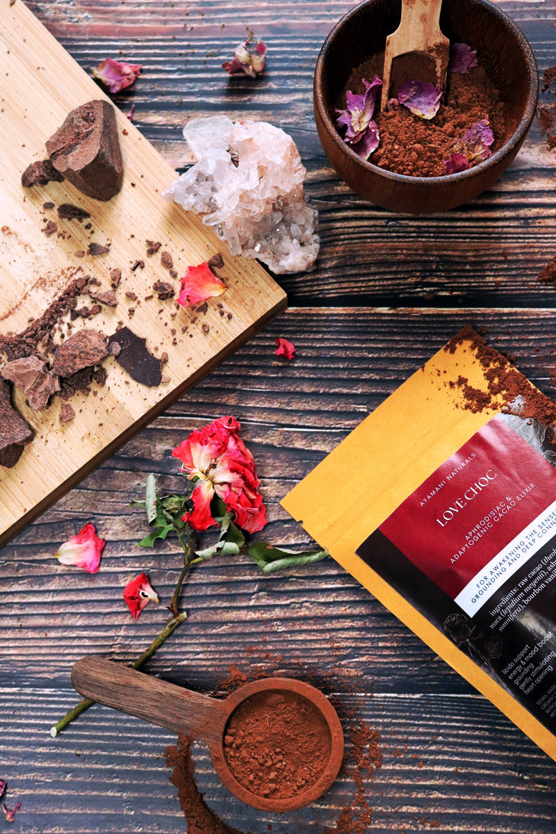 Aufnahme von oben von einer Packung „Love Choc Adaptogenic Cacao Elixir“, die diagonal auf einer dunklen Holzoberfläche platziert ist. Sie ist umgeben von Kakaopulver, einem Holzlöffel mit Pulver, einer kleinen Schale mit Pulver und Rosenblättern, Schokoladenstücken auf einem Schneidebrett, einer roten Rose und einem klaren Quarzkristall-Cluster.