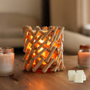 Ein rustikaler Kerzenhalter aus Holz, dessen helles, warmes Licht durch die Stäbchen scheint, umgeben von zwei kleinen Kerzengläsern und weißen Duftwachswürfeln.