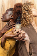 Zwei Frauen mit unterschiedlichen Hauttönen halten gemeinsam eine Flasche "PAAVANI AYURVEDA HERBAL HAIR OIL" mit geschlossenen Augen, ihre Hände verschränkt, was eine Verbindung und gemeinsames Wohlbefinden symbolisiert.