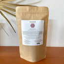 Ein großer brauner Papierbeutel mit Paavani Ayurveda Rosenblüten steht auf einer Holzoberfläche neben einem getrockneten Palmblatt.