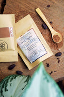 Zwei „Kaffee-Ersatz“-Superfood-Pulvermischungsbeutel liegen auf einer rustikalen Holzscheibe, begleitet von einem kleinen Holzlöffel mit Kakaopulver und verstreuten Kakaobohnen. Im Vordergrund befindet sich ein lebhaftes grün-weiß gesprenkeltes Blatt, das der Komposition eine frische, natürliche Note verleiht.