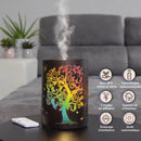 Ein dunkler elektrischer Aromatherapie-Diffusor mit einem regenbogenfarbenen Ausschnitt eines Baum-des-Lebens-Motivs, der leuchtet und Dampf abgibt, steht auf einem Tisch in einem Wohnzimmer, mit einer Grafik, die Merkmale wie BPA-freies Metall und Glas, Fernsteuerung, 2 Diffusionsmodi, Stimmungsbeleuchtung, Luftreinigung und automatische Abschaltung auflistet.