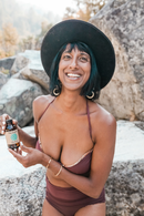 Eine lächelnde Frau mit dunklem Haar und schwarzem Hut steht draußen zwischen Felsen und hält eine Flasche PAAVANI Refreshing Mint Ölziehe-Öl. Sie trägt einen Nasenring und große Creolen.