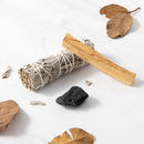 Ein Bündel weißer Salbei und ein Stock Palo Santo Holz liegen auf einer weißen Marmoroberfläche neben einem schwarzen Stein und getrockneten Blättern.
