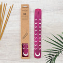 Rosa Räucherstäbchenhalter „Elm“ aus Holz mit silbernen Mondmotiven neben der Verpackung und Räucherstäbchen.