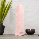 Eine hellrosa, zylindrische Yogamatten-Tasche mit dem vertikalen weißen „myoga“-Logo steht aufrecht auf einer Holzoberfläche, mit einer Topfpflanze und einer dekorativen Schale im Hintergrund.