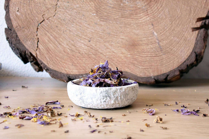 Eine Nahaufnahme von oben zeigt eine rustikale, hellgraue Schale, gefüllt mit einer lebhaften Mischung aus getrockneten violetten und goldenen Blütenblättern, vor einem unscharfen, hellbraunen Holzhintergrund. Die Texturen der Schale und der Blütenblätter sind deutlich erkennbar.