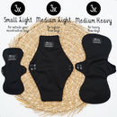 Drei Größen schwarzer Bamboozy wiederverwendbarer Binden (Small Light, Medium Light, Medium Heavy) auf einer gewebten Matte, jede mit Verwendungszweck (außerhalb der Menstruation, leichtere Tage, starke Tage) beschriftet.