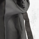 Eine Nahaufnahme des schwarzen Stoffes und des schwarzen Riemens einer Yogamattentasche mit einem kleinen gewebten Etikett mit dem Wort „Yoga“ (Teil des Logos) auf einem hellgrauen strukturierten Hintergrund.
