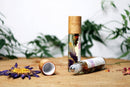 Eine Roll-on-Flasche mit Chakra-Öl und eine abgenommene Holzkappe liegen neben einer getrockneten blauen Lotusblüte mit gelbem Staubgefäß und verstreuten rosafarbenen Blütenblättern auf einer Holzoberfläche.