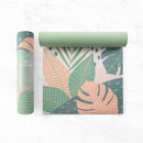 Eine gerollte und teilweise ausgerollte hellgrüne Yogamatte mit tropischem Palmenblatt-Muster in Pfirsich, Grün und Dunkelblau neben der passenden Markenverpackung.