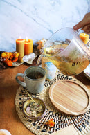 Eine Hand gießt Kräutertee aus einer Glaskanne in eine Tasse. Auf dem Tisch stehen Kerzen, getrocknete Blumen und eine kleine Schale mit getrockneten Kräutern.