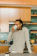 Ein Mann mit geflochtenen Haaren steht in einer modernen Küche und trinkt aus einer Keramikschale.