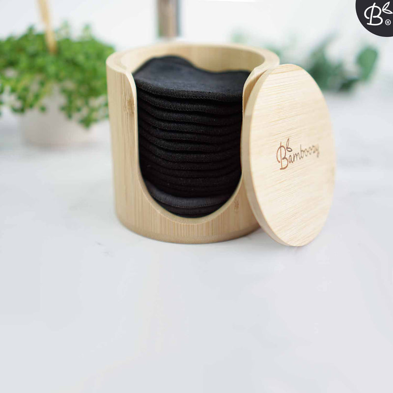 Ein Bambushalter für wiederverwendbare Wattepads, gefüllt mit schwarzen Pads, steht auf einer weißen Arbeitsplatte mit einer unscharfen Grünpflanze im Hintergrund. Der Deckel mit "Bamboozy"-Logo ist leicht geöffnet.