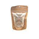  Eine transparente Plastikverpackung mit braunem Boden, die ein Paavani Augenwaschbecher-Set enthält, bestehend aus einem Glas-Augenbecher und einem kleinen Stoffbeutel, auf einem weißen Hintergrund.