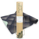 Eine teilweise ausgerollte Myga Yogamatte aus veganem Wildleder auf Gummi mit einem dunklen, 3D-karierten oder rautenförmigen Muster, die diagonal hinter ihrer hohen, gebrandeten braunen Kartontragrolle liegt.