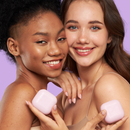 Zwei lächelnde Frauen mit nackten Schultern halten jeweils ein pastellrosa Stück Seife vor einem hellvioletten Hintergrund. Die Stimmung ist fröhlich und frisch.