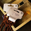 Rudraksha-Mala-Gebetskette und zwei weiße Produktetiketten, die teilweise in einer Holzkiste liegen, mit einer goldenen Quaste in der Ecke.