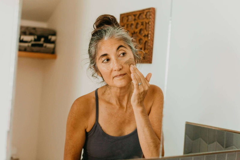  Eine Frau mit teilweise aufgetragener Gesichtsmaske blickt in einen Spiegel und hält ihre rechte Hand an ihre Wange, als würde sie die Maske glätten, ihr Haar ist zu einem Dutt gebunden.